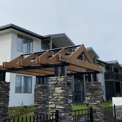 The modern two-story building boasts white walls and balconies, complemented by a hand brushed wooden pergola with stone pillars. It's framed by a black metal fence and lush greenery.