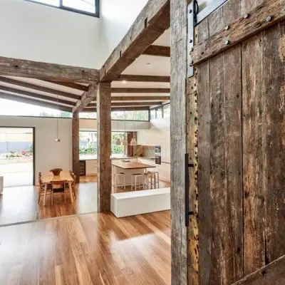 Open-plan interior with wooden beams and structural beams accentuating the space. A large rustic sliding door complements the wooden flooring. Dining table, chairs, and kitchen island are visible, offering a view of the garden through expansive windows.