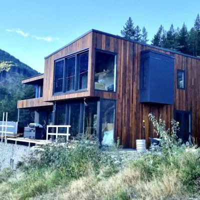 The modern two-story wooden house, sawn with oil for a refined finish, features large windows that embrace the serene hillside view surrounded by trees and mountains.