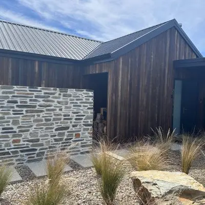 Modern house exterior with wooden siding treated in clear oil, featuring a sleek metal roof. A stone wall partially encloses a courtyard adorned with dry landscaping elements like sawn rocks and grasses under a clear sky.