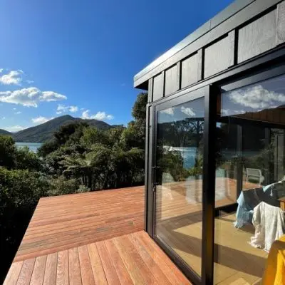 Modern wooden deck and glass-walled building with view of lush greenery, mountains, and a body of water under a clear blue sky.