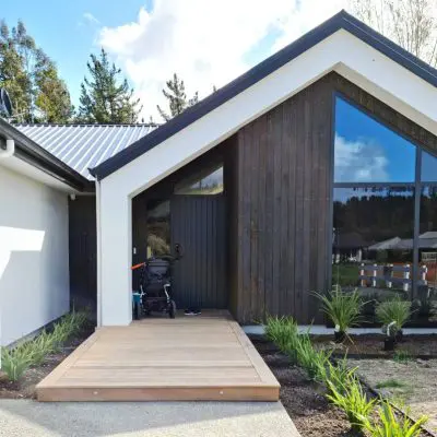 Modern house exterior with dark wood and white walls, featuring large windows with a subtle smokey tint. A wooden deck surrounded by lush plants enhances the serene vibe, while a stroller is parked near the entrance.