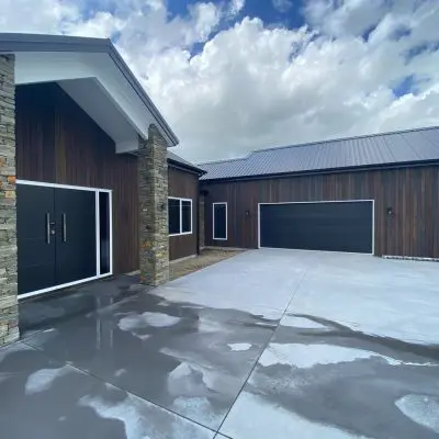 A modern house with dark wood cladding and stone columns, featuring sawn surfaces and large black garage doors. The wet concrete driveway, with a lightly brushed finish, glistens under a cloudy sky.