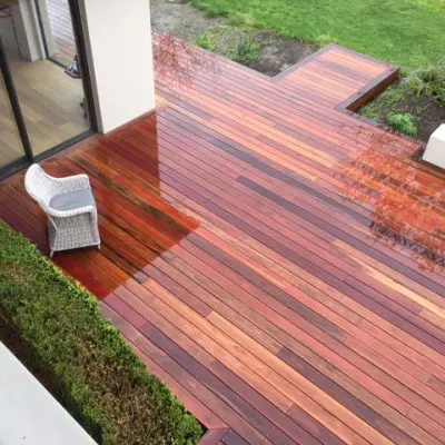 A wet wooden deck with a single wicker chair, surrounded by greenery and attached to a modern building with glass sliding doors.