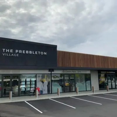 The exterior view of the shopping center showcases a striking black and hardwood facade, featuring "The Prebbleton Village" sign. Thoughtfully designed landscaping complements the scene, while empty parking spaces are visible in the foreground.