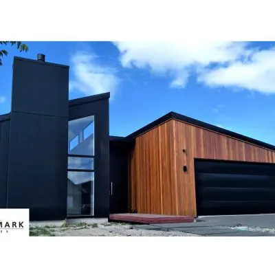 Modern house with a black and wood exterior, featuring large windows and a slanted roof. Blue sky with clouds in the background. Landmark Homes logo in the corner.