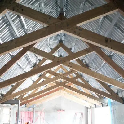 Hardwood trusses diligently support a corrugated metal roof in the building under construction, displaying visible bolts and beams.