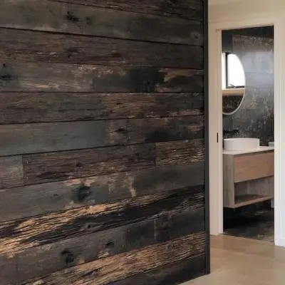 A room with a weathered, dark wooden accent wall on the left and a partial view of a bathroom through an open door on the right. The bathroom features a round mirror and sink, deeply enhancing the rustic charm.