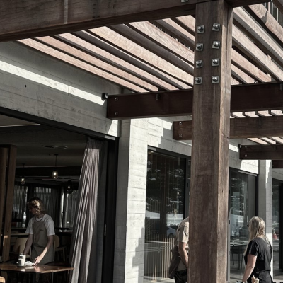 Wooden pergola structure outside a modern building with large glass windows, a man standing by a table, and a woman walking past. Cloudy sky in the background.