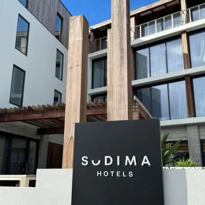 The exterior view of Sudima Hotels showcases a modern facade design, featuring large windows and wooden accents. A black sign with the hotel name is prominently displayed in the foreground, highlighting its stylish exterior cladding.