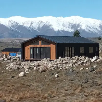 A modern wooden cabin crafted from Silvertop Ash boasts a sleek black roof, perched on rocky terrain. Snow-capped mountains paint the backdrop under a pristine blue sky.