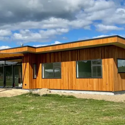 Modern single-story house with a timber exterior, large windows, and a covered patio, set against a backdrop of grass and a partly cloudy sky.