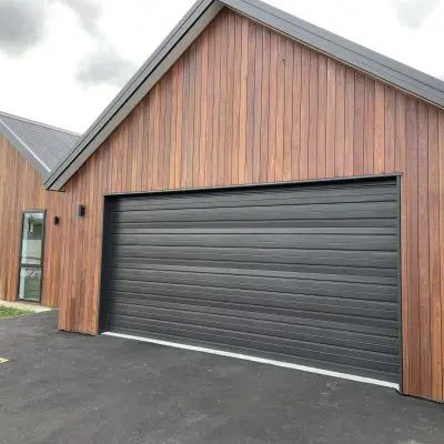 Modern house with wooden siding crafted from Queensland Spotted Gum, and a black, paneled garage door. It features two narrow windows on the left side adjacent to a paved driveway.