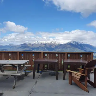 Outdoor deck with wooden tables and chairs, overlooking snow-capped mountains and a cloudy sky.