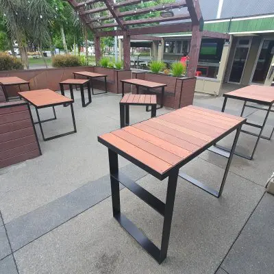 Outdoor seating area with multiple wooden high tables and greenery, under a wooden pergola near a building entrance.