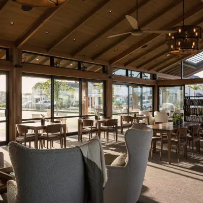 A spacious, sunlit lounge with wooden ceiling, armchairs, and tables. A bookshelf is on the right, and large windows offer views of greenery outside.