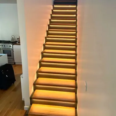 A staircase with illuminated steps leading upwards in a modern interior, adjacent to a kitchen area with a stove and countertop.