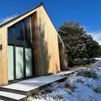 A modern wooden house with weathered cladding and large windows stands serenely amidst snow-covered ground and trees, all under a clear blue sky.