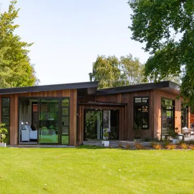 Modern wooden house with large windows, surrounded by green lawn and trees on a sunny day.