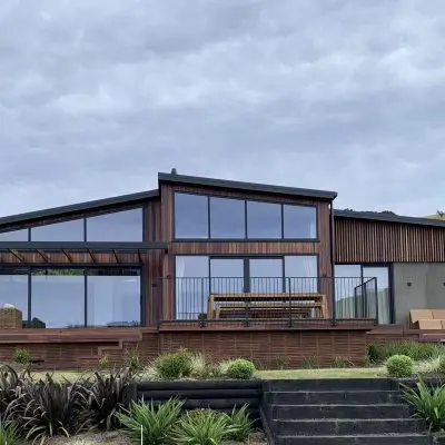 Modern house with large windows, wooden and concrete elements, surrounded by greenery under a cloudy sky. Steps lead up to the entrance.