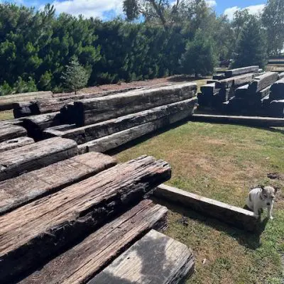 Large, aged wooden beams stacked in an outdoor area with grass and gravel pathways. A small dog stands nearby, adding a touch of charm. Trees in their natural state frame the scene under a partly cloudy sky.