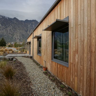 The wooden house, featuring weathered cladding and large windows, is set against a mountainous backdrop. A gravel path lined with plants leads from the foreground to the home, adding to its rustic charm.