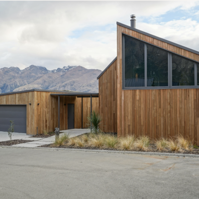 Modern wooden house with large windows and sloped roof, featuring weathered cladding that seamlessly blends with the mountain backdrop. Includes a driveway, garage, and minimal landscaping adorned with grasses and small plants.