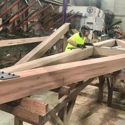 Two workers in safety gear construct a large wooden structure inside a workshop, surrounded by tools and machinery.