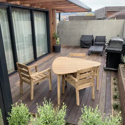 Wooden patio with a triangular table and two chairs, a grill on the right, two lounge chairs with covers, and potted plants, against a house with sliding glass doors and wooden exterior.