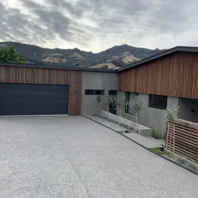 Modern house with wood paneling and large garage, set against a backdrop of hills and a cloudy sky. A concrete driveway leads to the entrance.