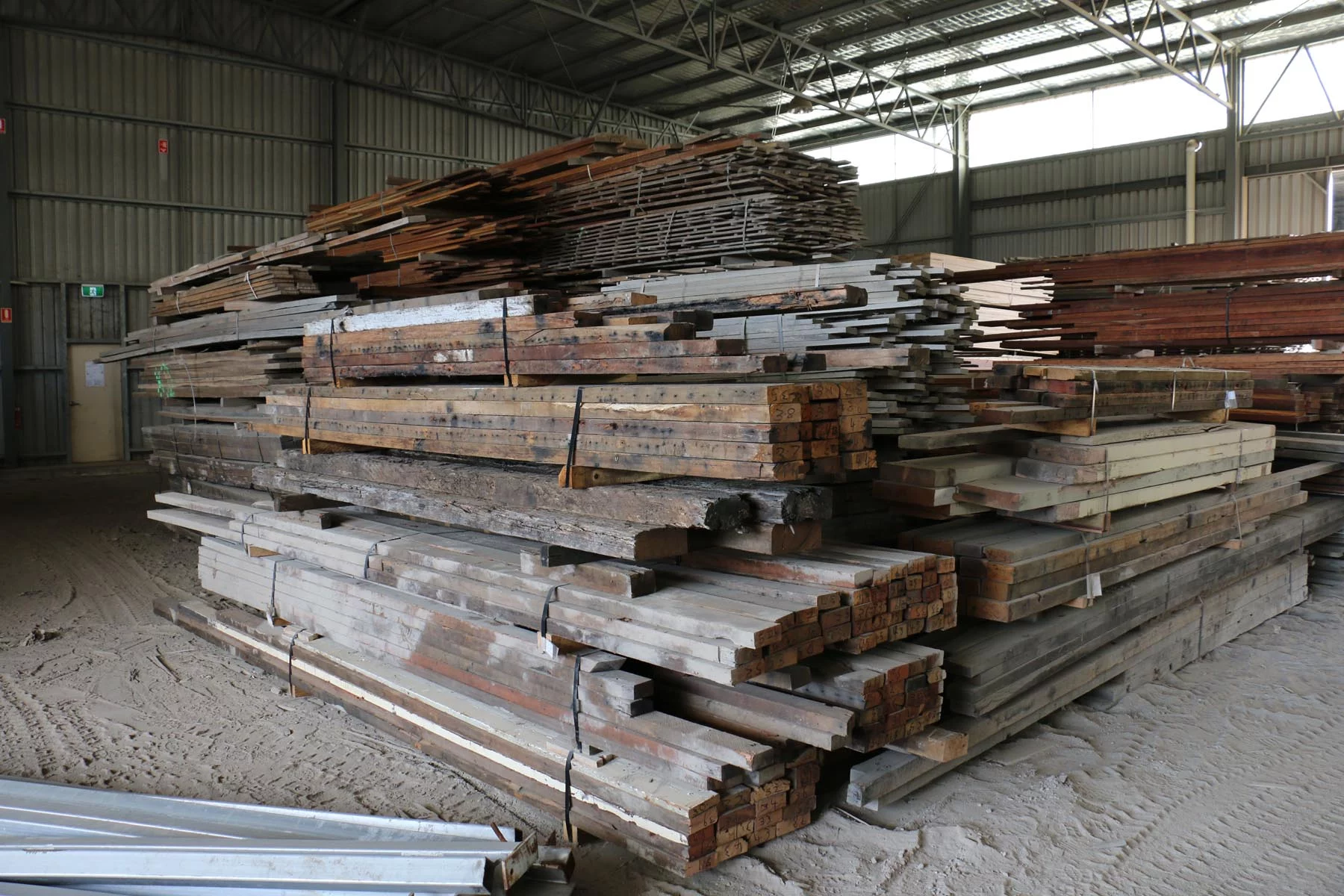 Stacks of assorted wooden planks and boards in a large warehouse with a metal roof.