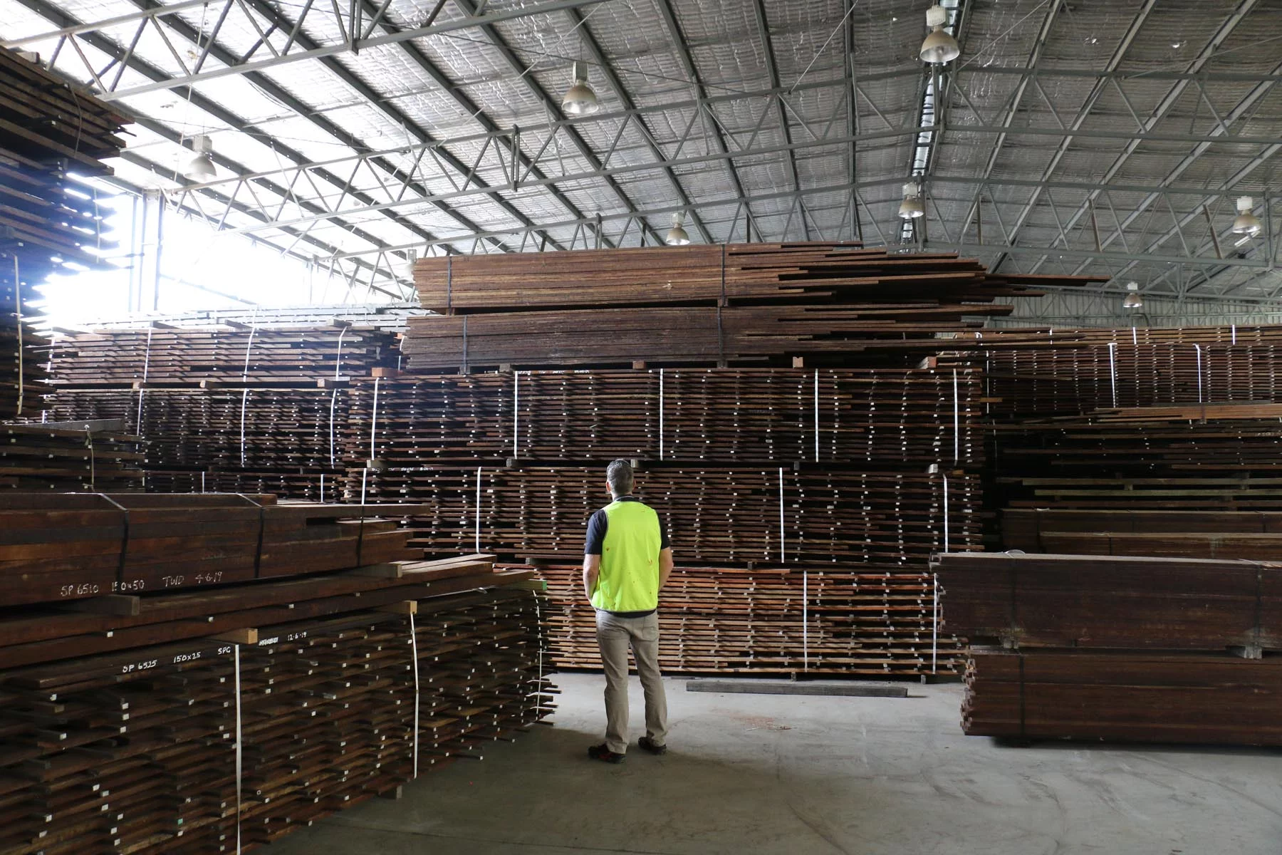 A person in a yellow vest stands in a warehouse, facing large stacks of neatly piled wooden planks.