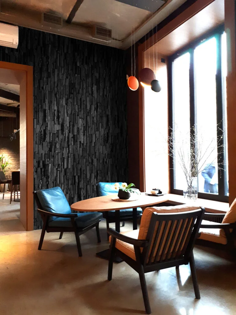 Stylish room with wooden furniture, a round table, blue chairs, and a textured dark accent wall enhanced by elegant interior cladding. Large windows provide natural light, complemented by hanging lights and minimalist decor.