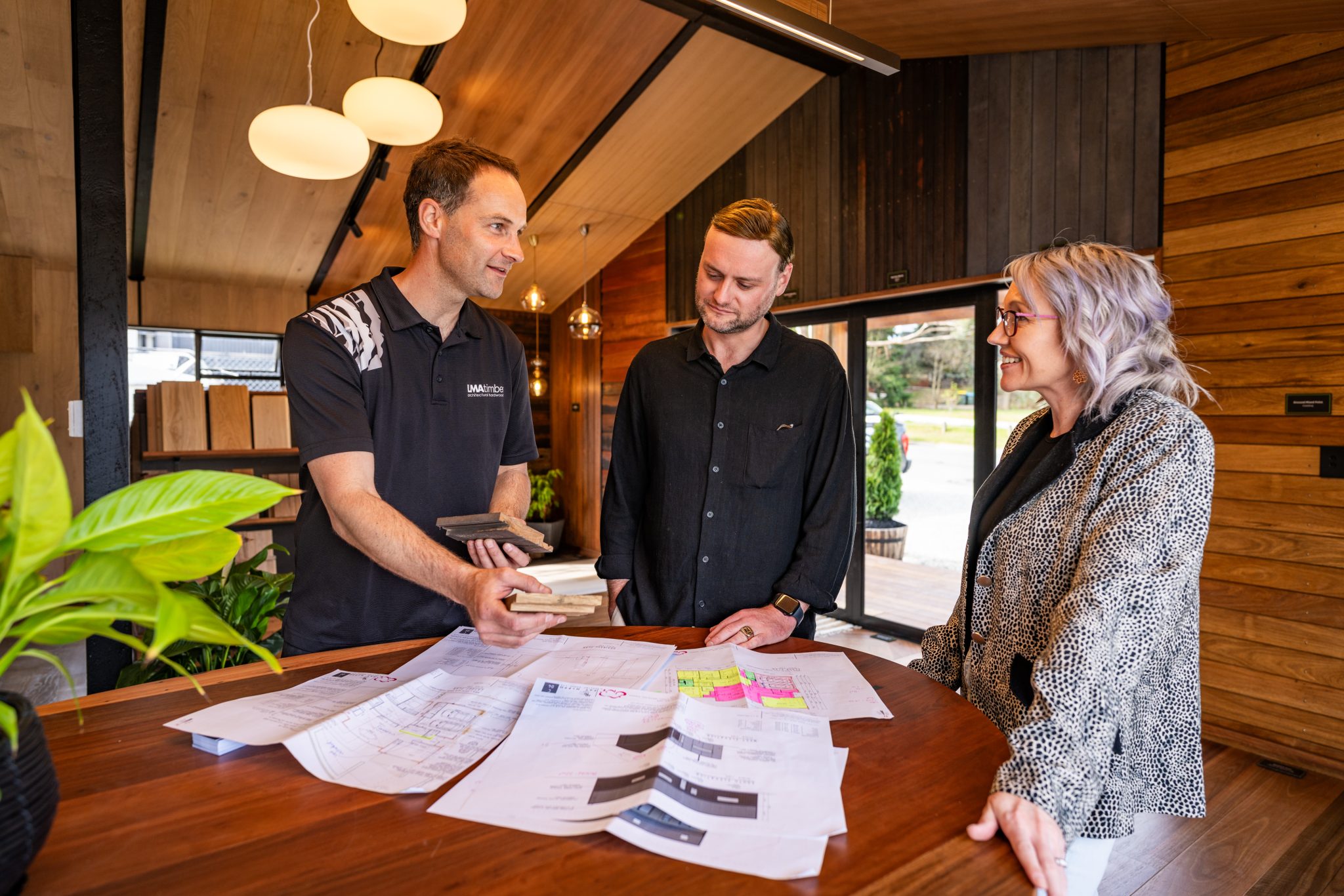 Three people in Christchurch are discussing house designs at a wooden table. Architectural plans and samples are spread out, while the two standing individuals pay attention to someone presenting materials.