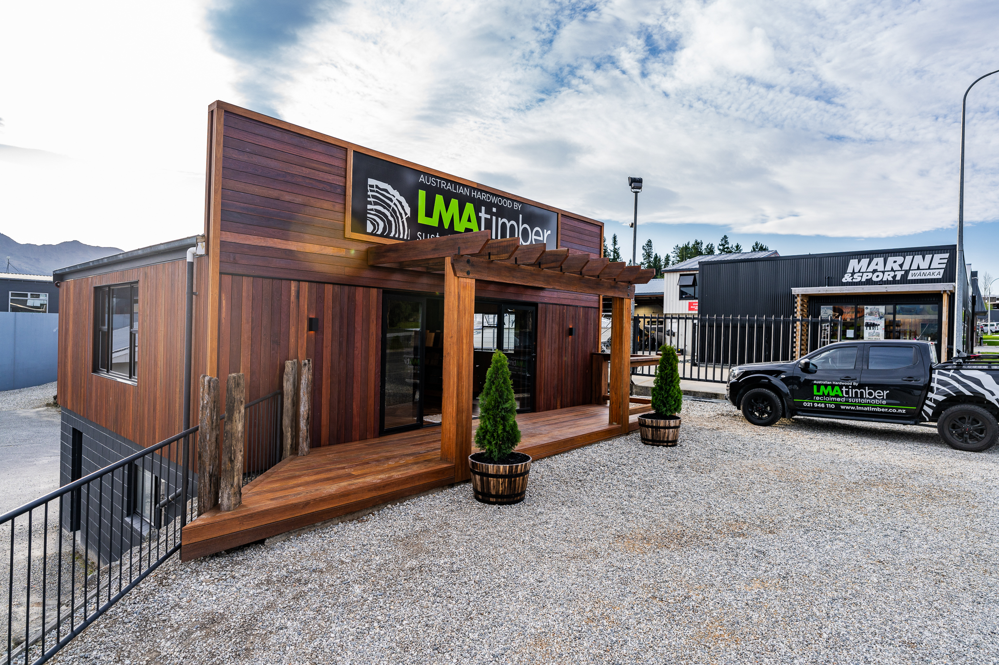 A wooden building with "LMA Timber" signage stands proudly, flanked by two potted plants at the entrance. Nearby, a branded pickup truck is parked by the roadside. In the background, a building displaying "Marine & Sport" hints at Tauranga's vibrant community.